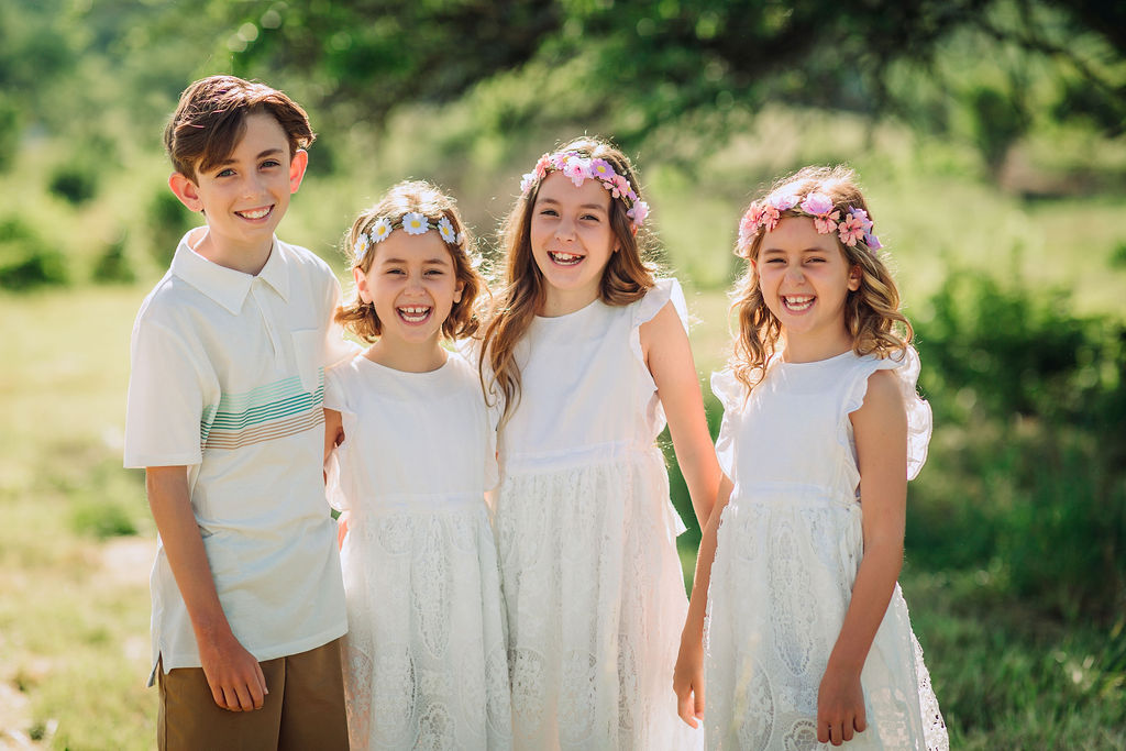Four siblings laugh and play in a park with the three sisters wearing matching white dresses and floral headbands moutain ridge pediatrics
