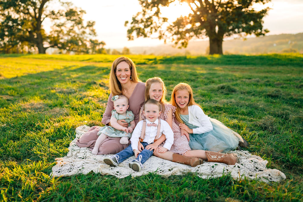 A mother in a pink dress sits on a lace picnic blanket with her three daughters and one son in a grassy field at sunset