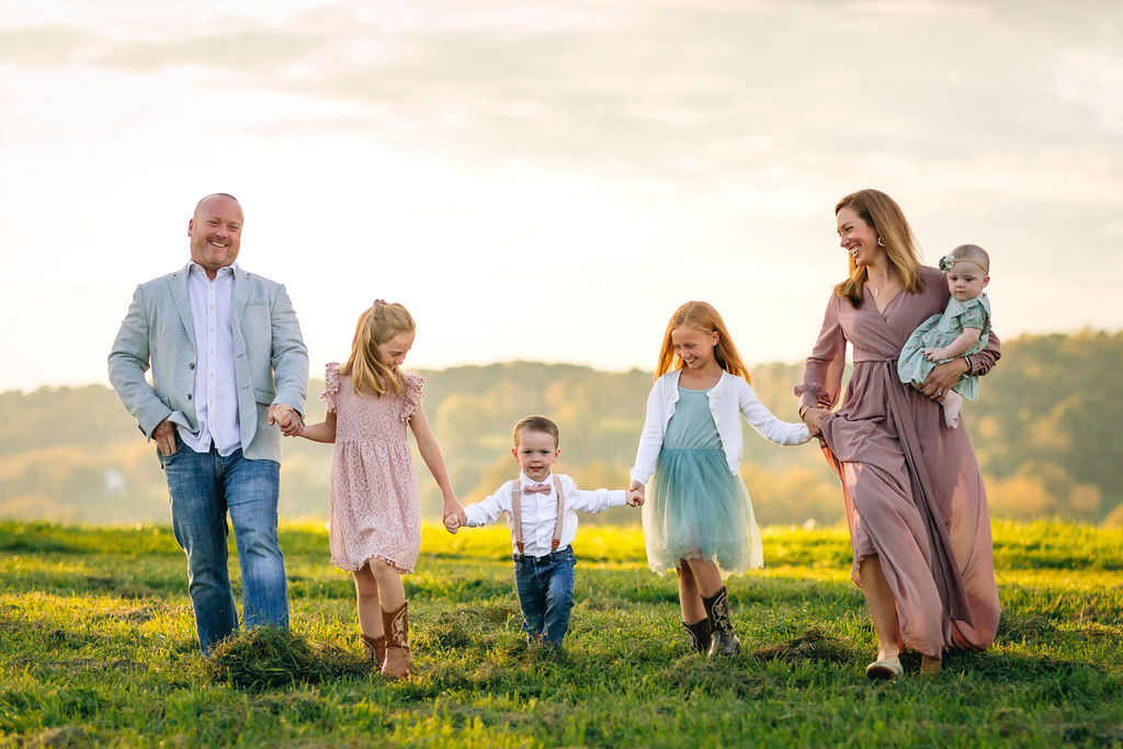 A mother and father walk through a grassy field while holding hands with three of their children and mom carries the toddler daughter on her hip caitlin batchelor dentistry