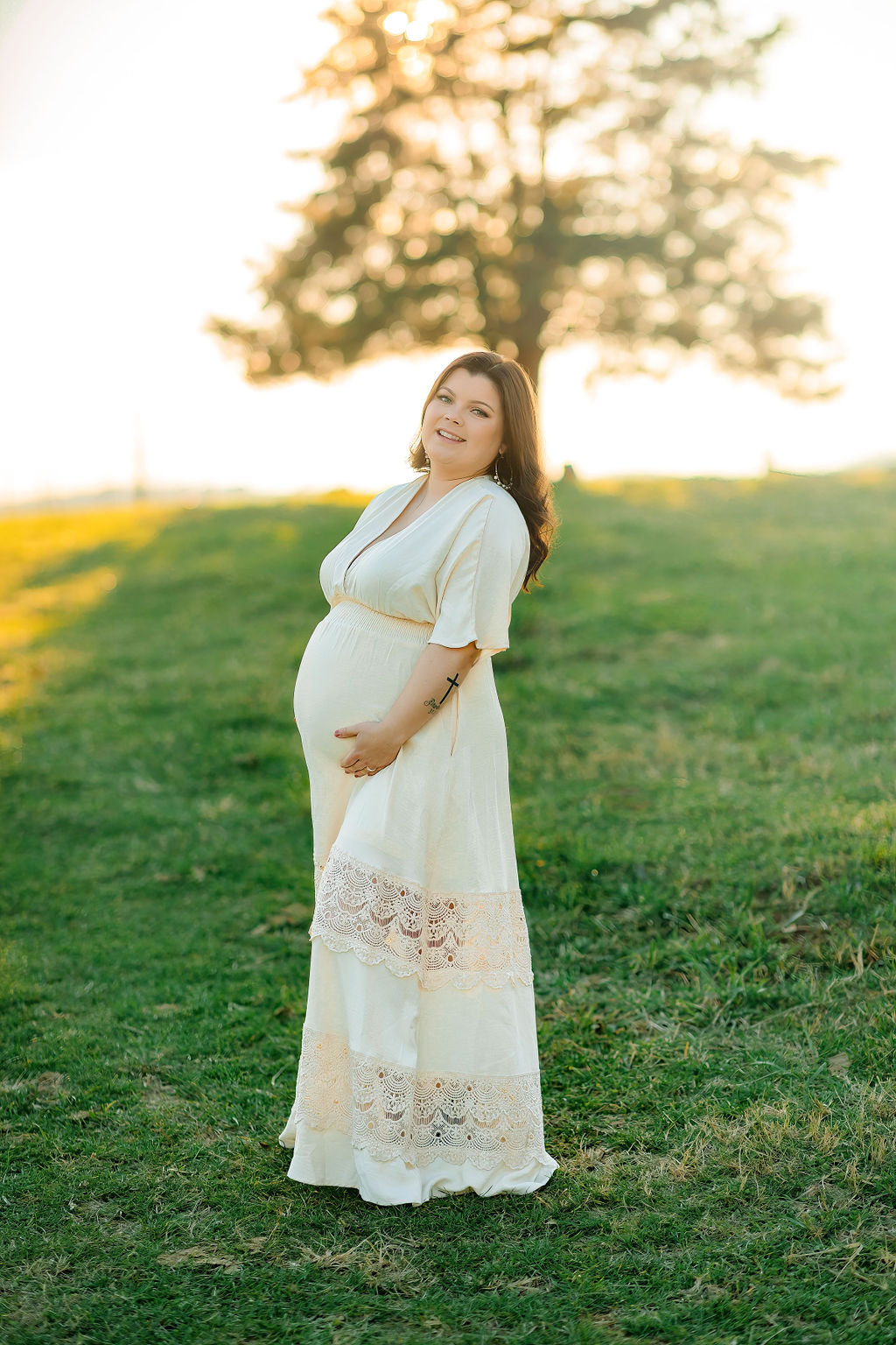 A mother to be stands in a grassy field holding her bump in a white dress