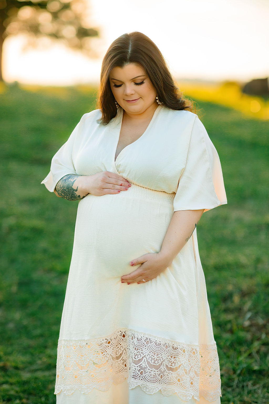 A mother to be in a white and lace maternity dress stands in a green field looking down at her bump cedar stones wellness spa