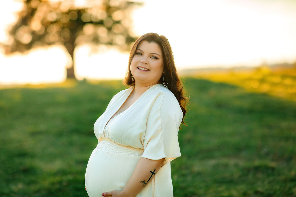 A mom to be holds her bump while standing in a green park at sunset cedar stones wellness spa