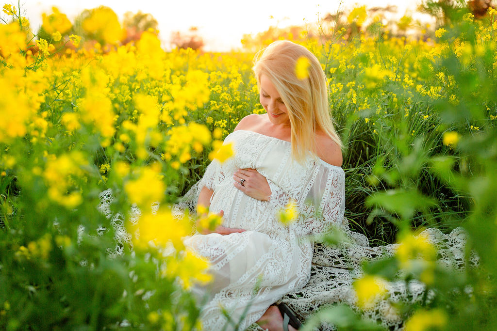 A mother to be in a white lace maternity dress sits in a bed of yellow wild flowers center for advanced gynecology