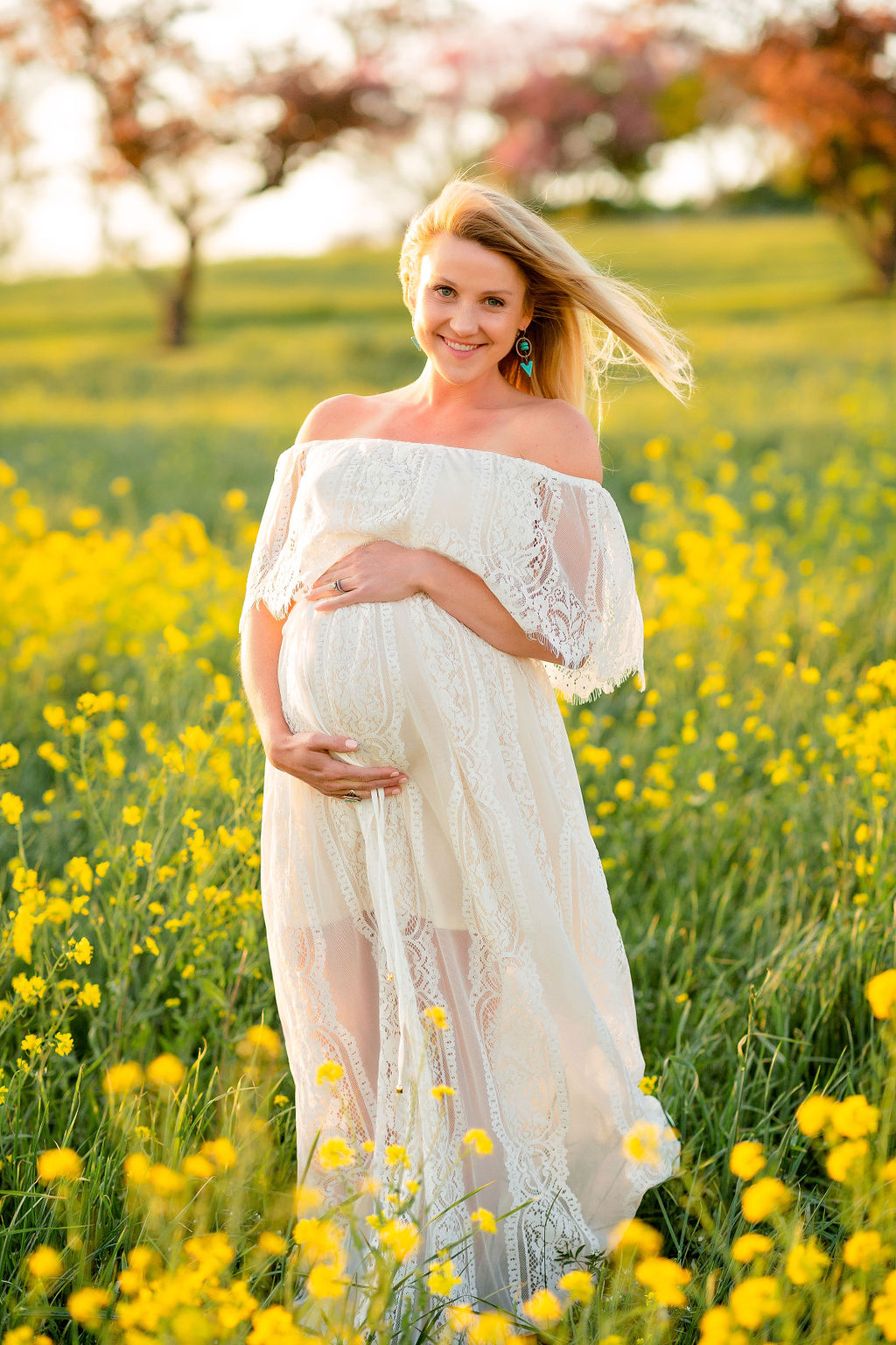 A mom to be in a lace maternity dress stands in a field of yellow wildflowers while holding her bump