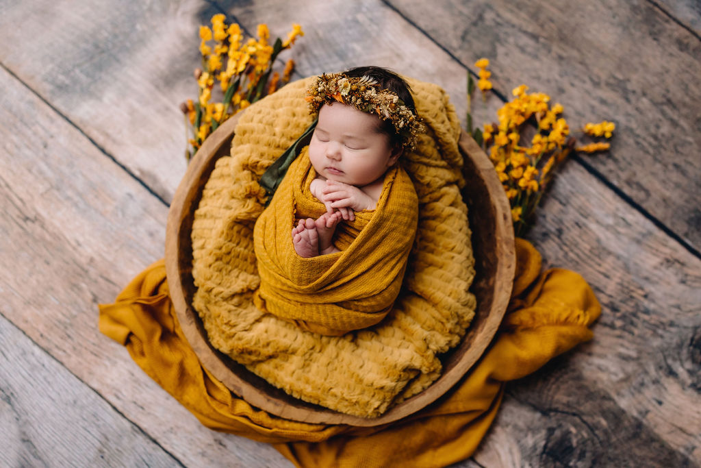 A newborn baby sleeps in a swaddle and blanket inside a wooden bucket with yellow flowers around her charlottesville daycare