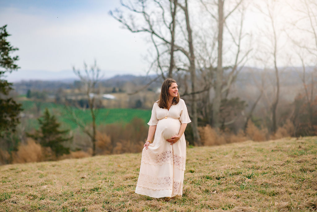 A mother to be in a lace maternity dress walks through a field holding her dress and her bump harrisonburg obgyn