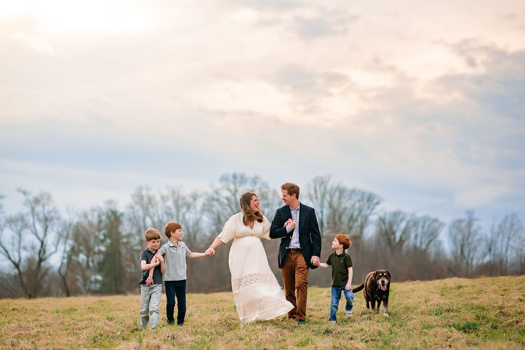 A pregnant mother walks hand in hand with her husband and three older sons through a field while their brown dog walks with them harrisonburg obgyn