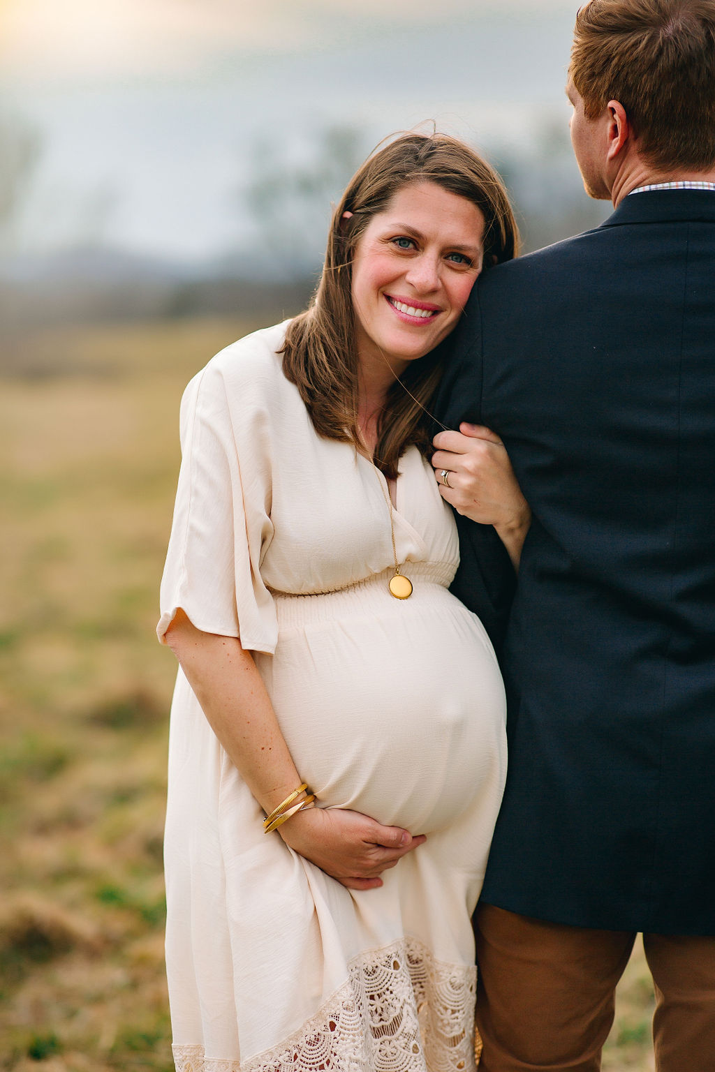 A mom to be holds on to and leans on the arm of her husband while wearing a cream maternity dress