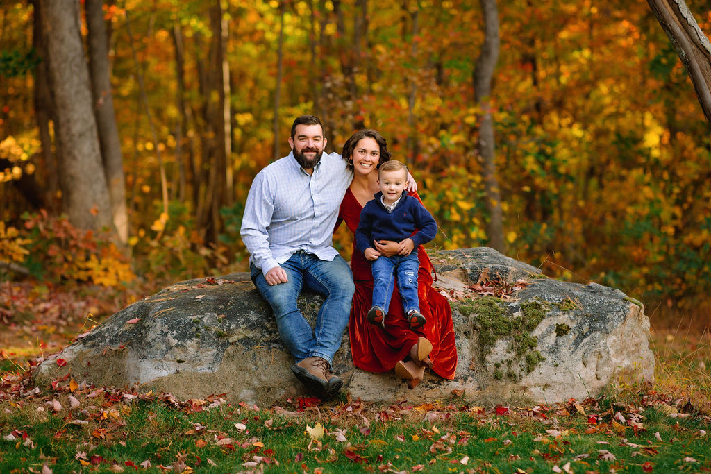 A family of three sit on a large boulder in a forest