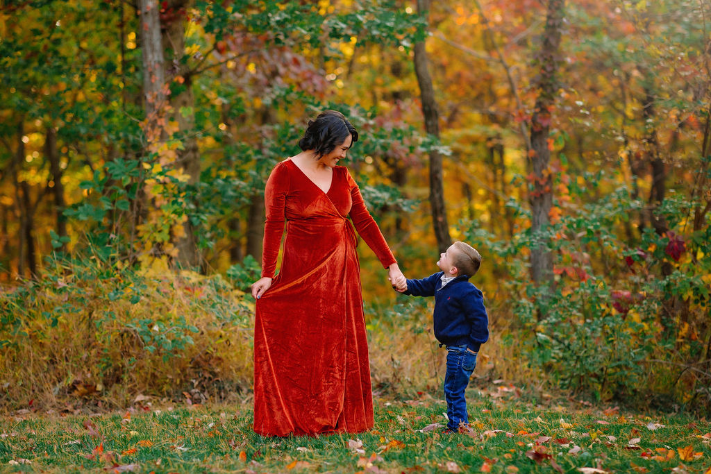 A mother in a red dress walks and plays with her young son in jeans and blue sweater through a park field in fall harrisonburg pediatric dentistry