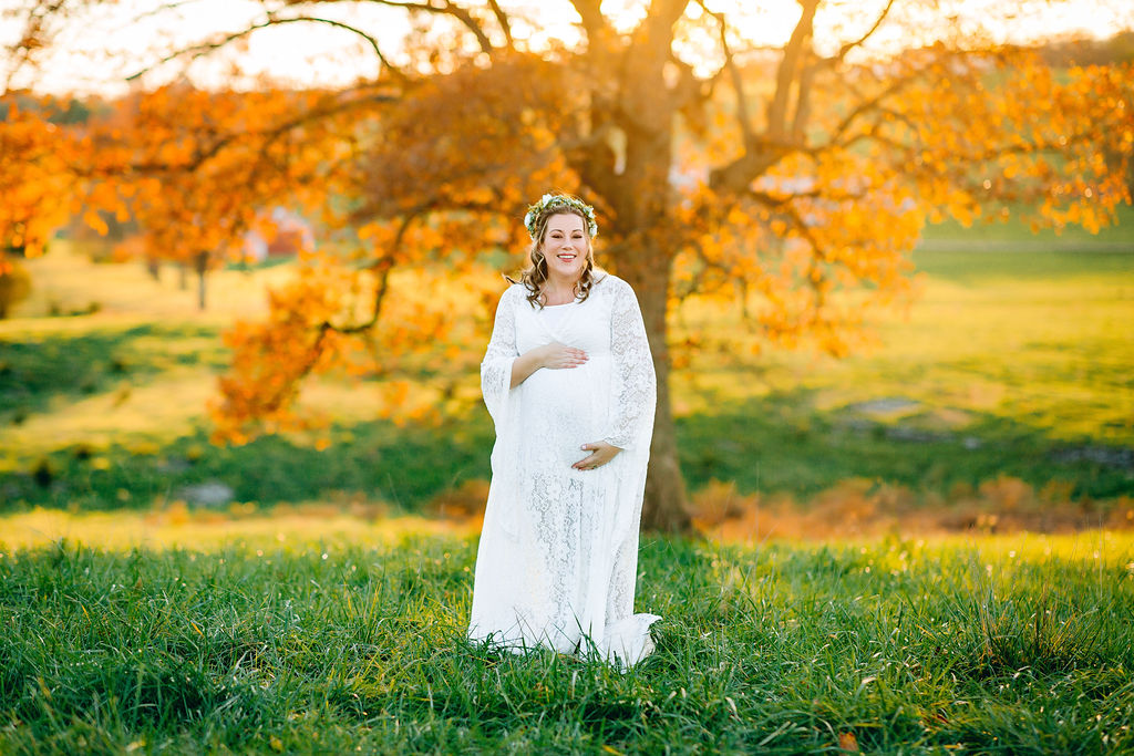 A mom to be walks through a grassy green field at sunset while holding her bump in a white lace dress