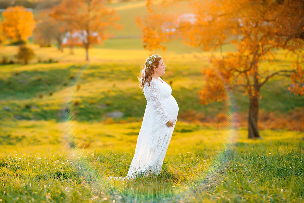 A mother to be stands in a white lace dress holding her bump in a field at sunset kline chiropractic