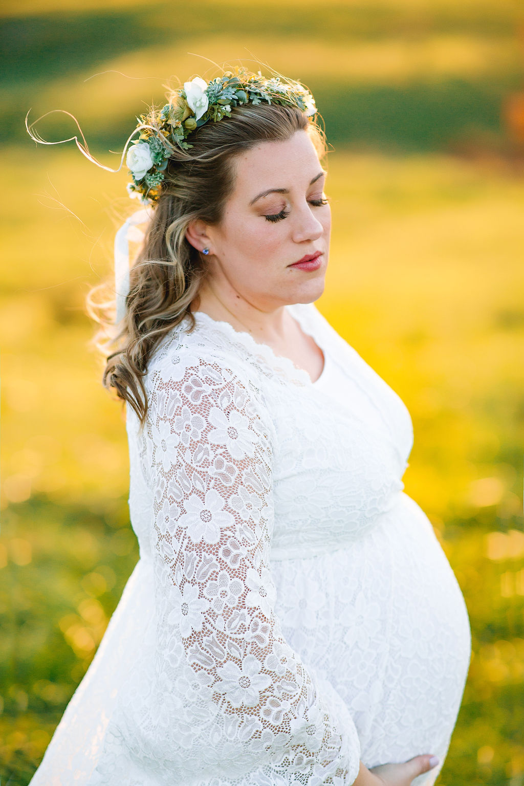 A mother to be stands in a grassy field in a white lace dress and floral headpiece kline chiropractic