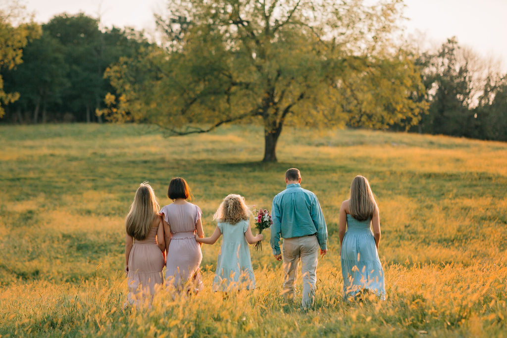 A family of five with three daughters walk through a field of tall grass while holding hands at sunset liberty mills farm
