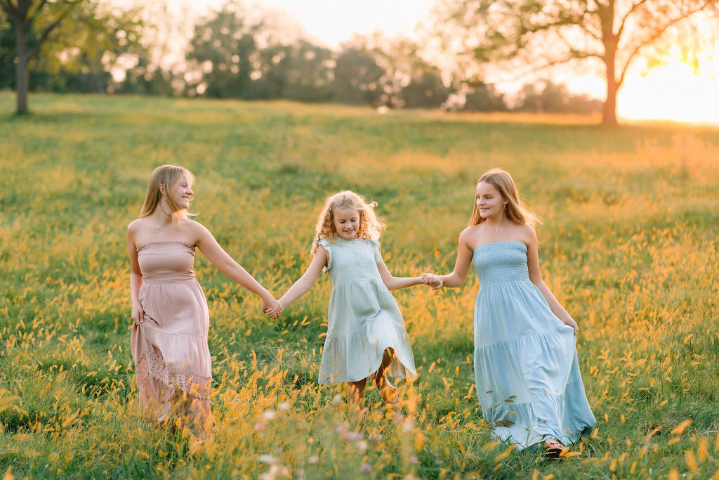Three sisters in along dress walk through a field of tall grass holding hands liberty mills farm