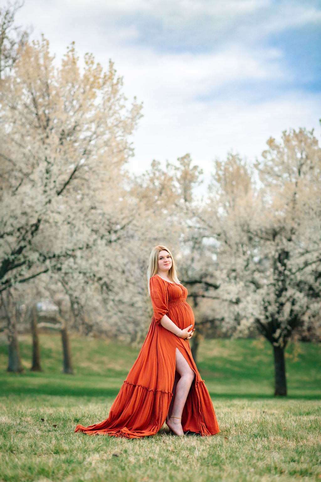 A mother to be in a red maternity gown stands in a field surrounded by white blooming trees shenandoah chiropractic
