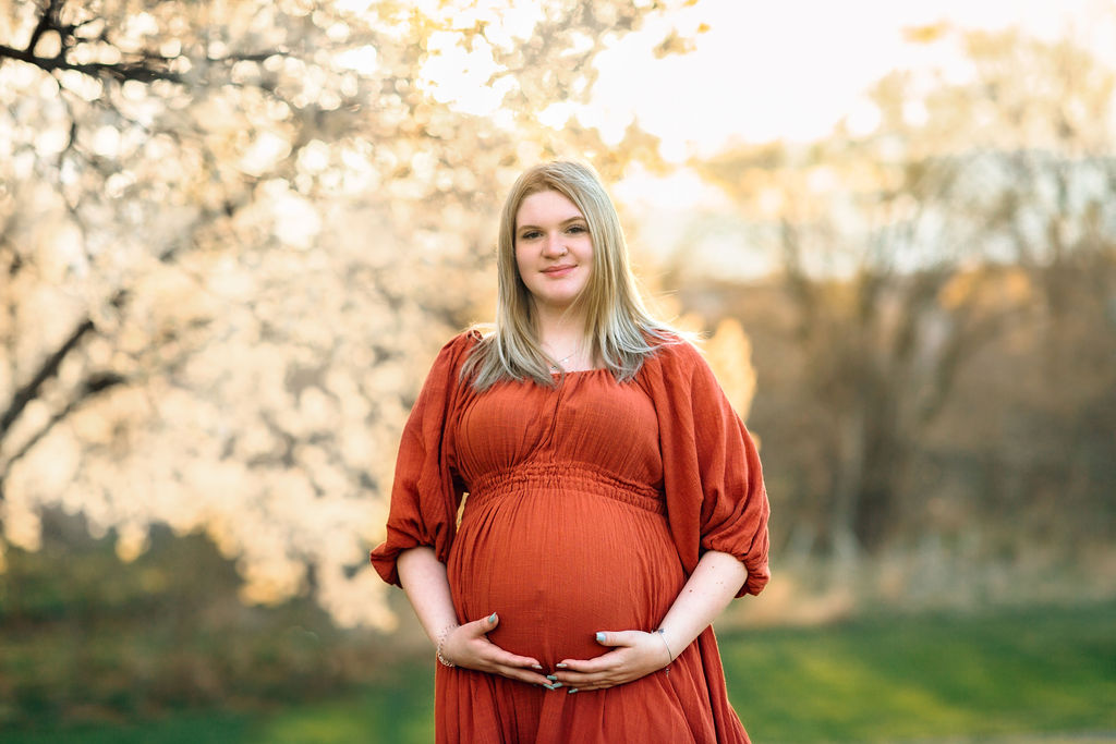 A mother to bee stands under white blooming trees in a red maternity gown shenandoah chiropractic
