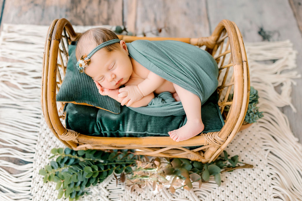 A newborn baby in a green blanket sleeps on its side in a wicker bed