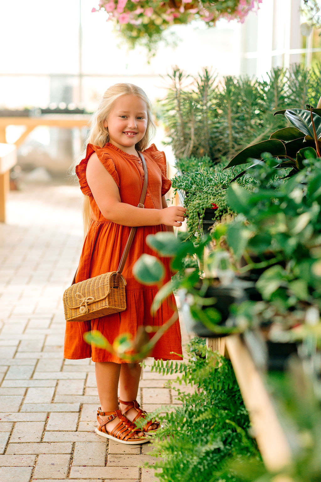 A young girl wearing a red dress browses some plants inside a bright greenhouse