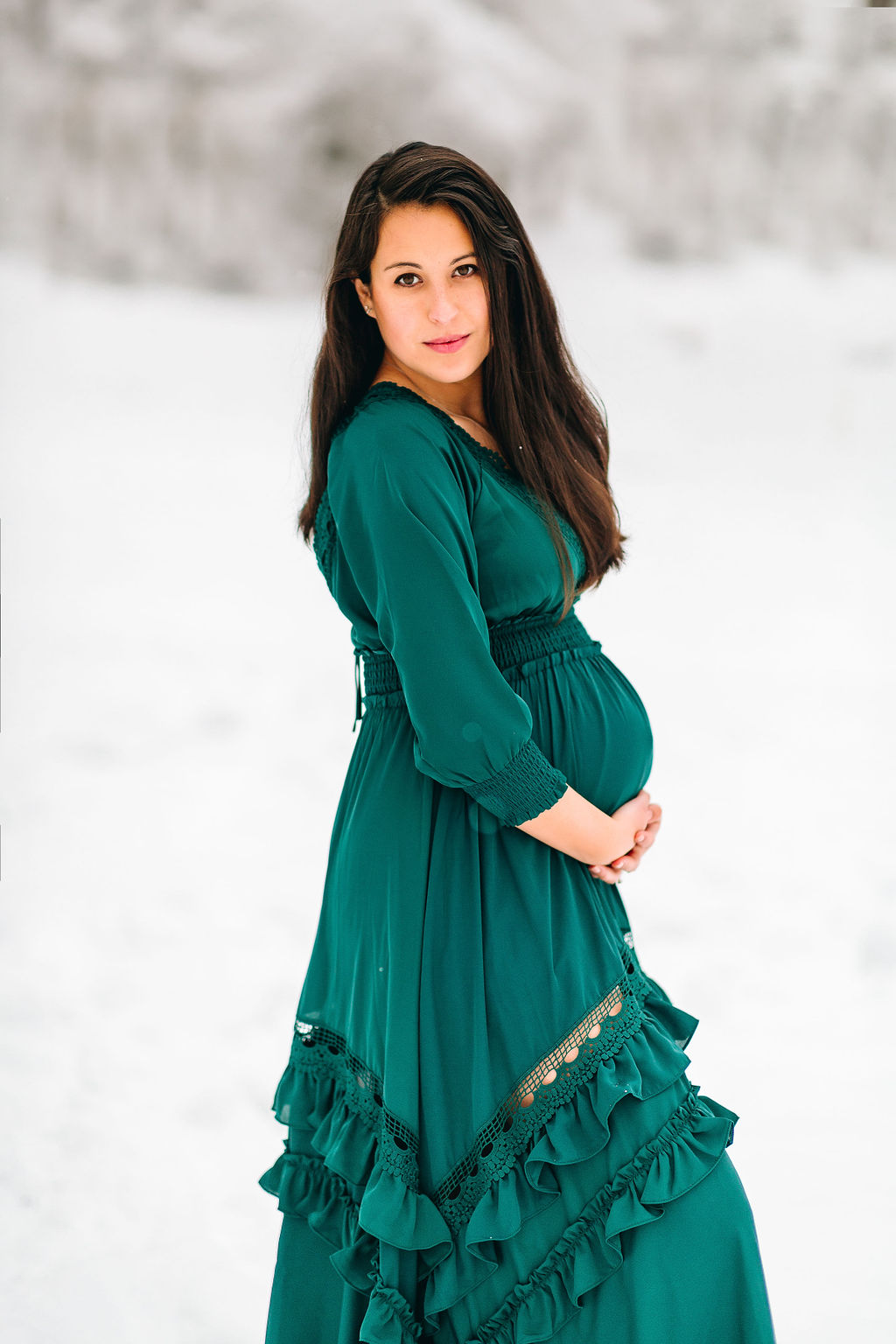A mother-to-be in a teal maternity dress stands in a snowy forest while holding her bump women in motion