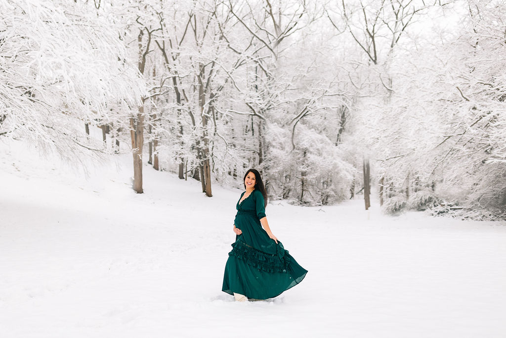 A mother to be walks and plays with her teal maternity dress in a snowy forest