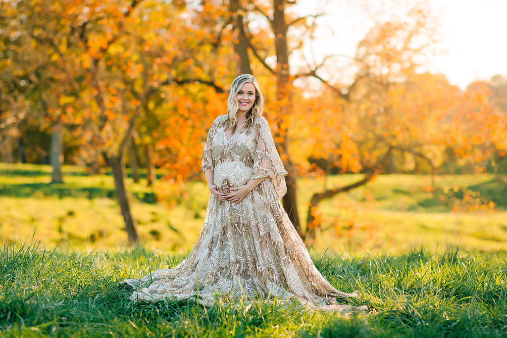 A mother to be in an ornate maternity gown stands in a field of green grass at sunset