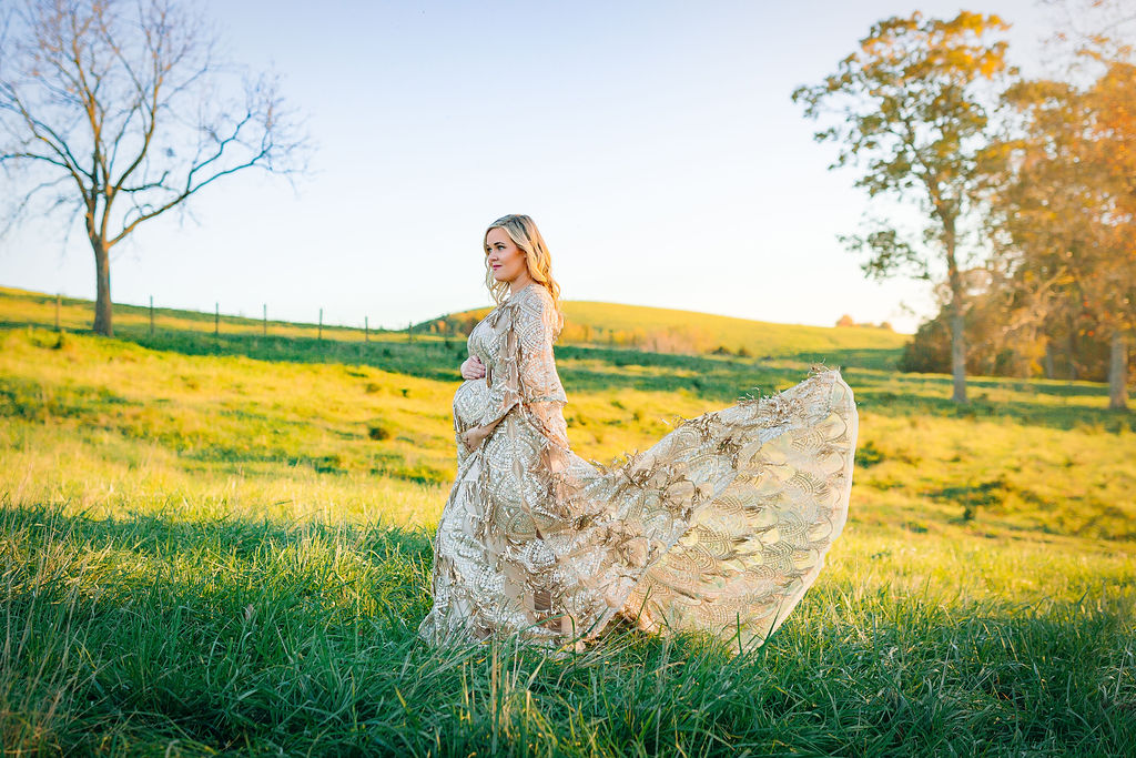 A mom to be stands in a field of grass in an ornate maternity gown that is flowing behind her Charlottesville ivf