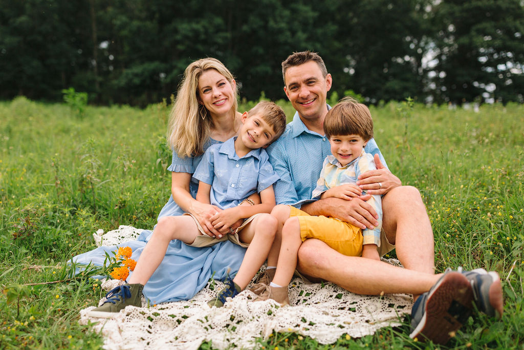 A mother and father sit on a lace picnic blanket in a field while their two sons sit in their laps charlottesville preschools