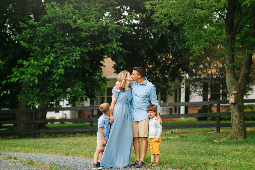 A mother and father kiss while standing in a green yard with their two young sons by their sides