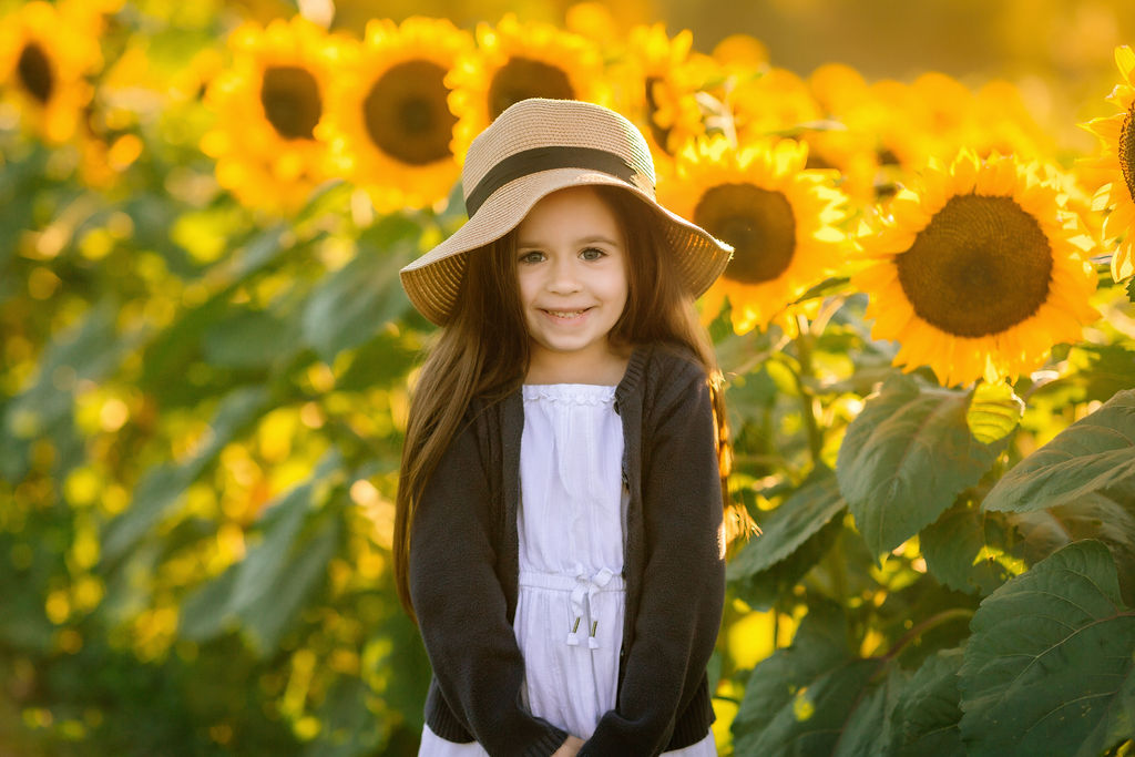 A young girl stands in a sunflower field at sunset while wearing a large hat