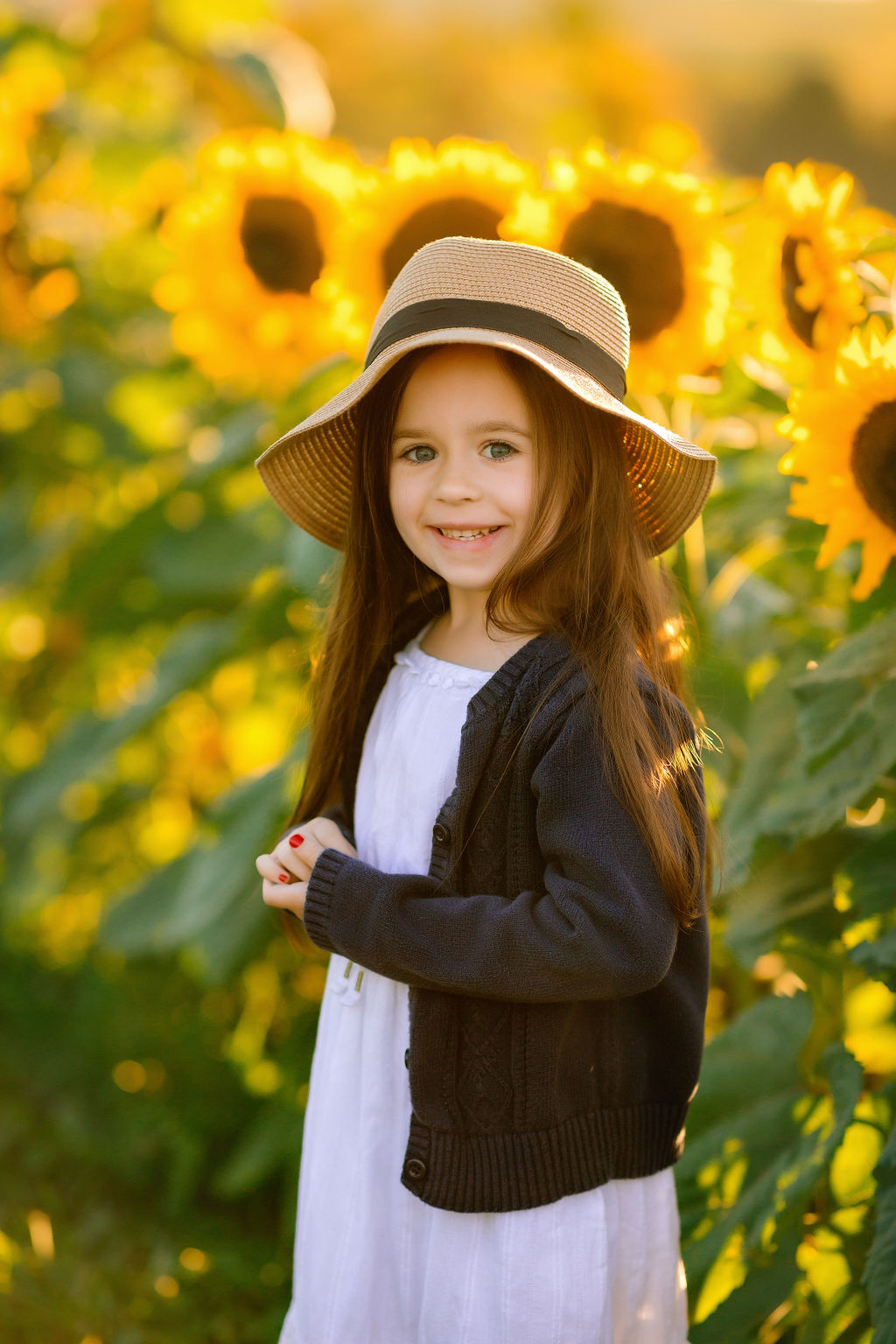 A young girl in a big woven hat, black sweater and white dress stands in a field of sunflowers inmotion school of dance