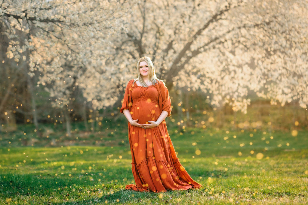 A mother to be stands in a field of blooming trees holding her bump nine moons midwifery