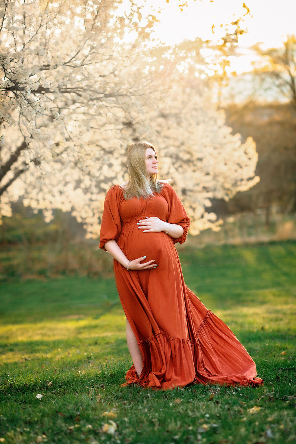 A mother to be stands in a flowing red maternity dress under a blooming tree at sunset nine moons midwifery