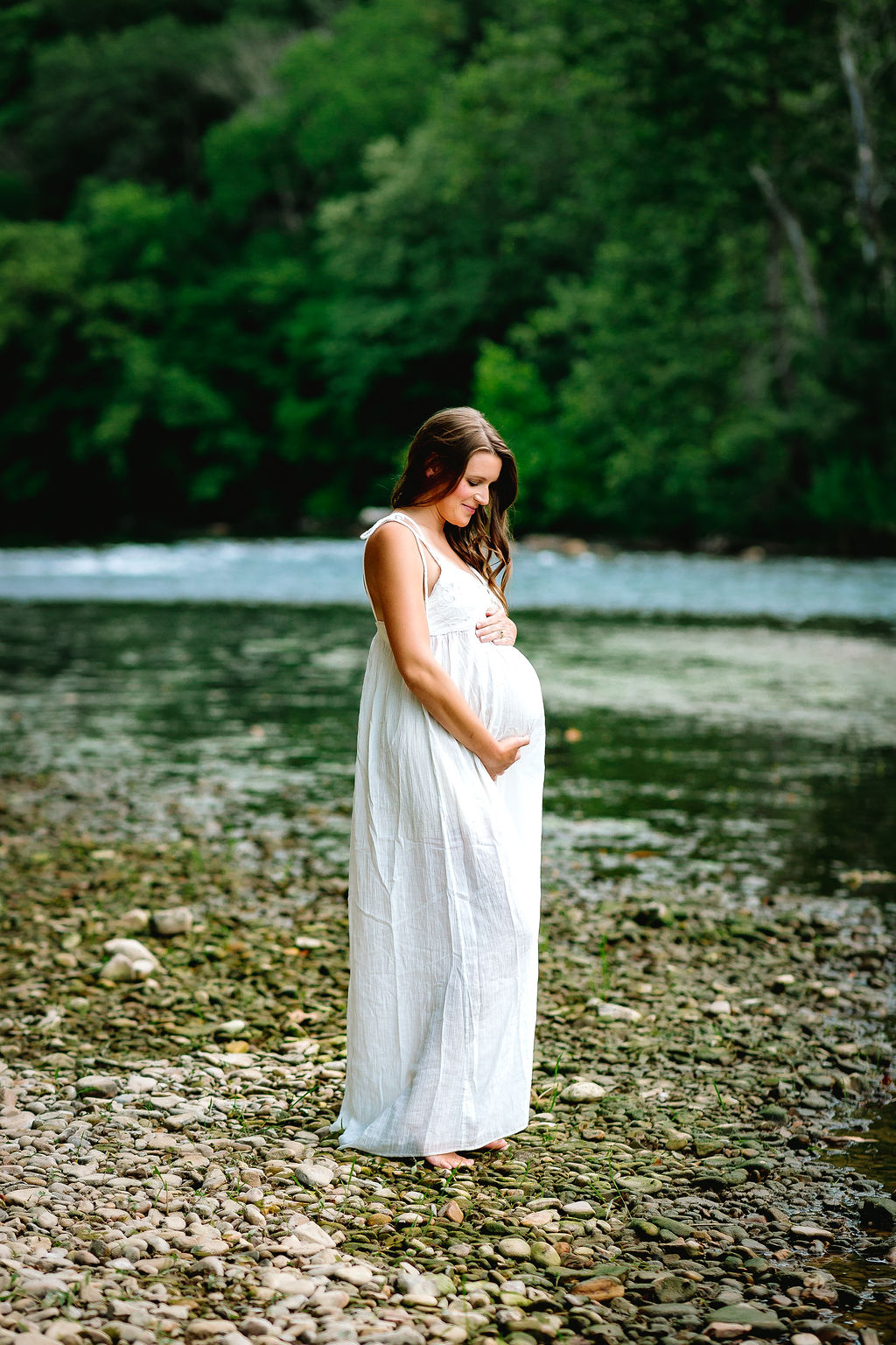 A mom to be stands on the edge of a river looking down at her bump in a white maternity gown