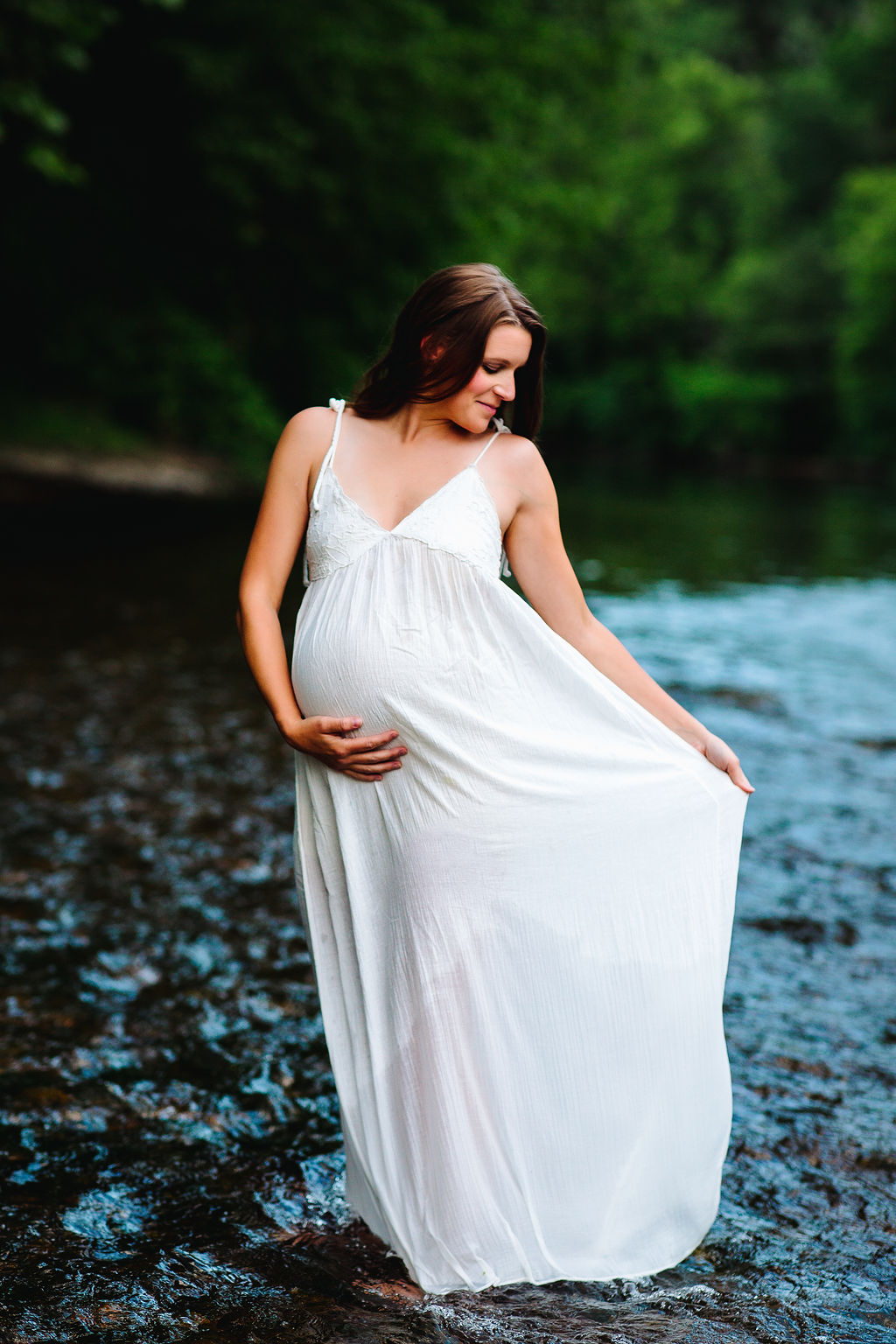 A mother to be stands in a river holding her bump and her white maternity dress obgyn stanton va