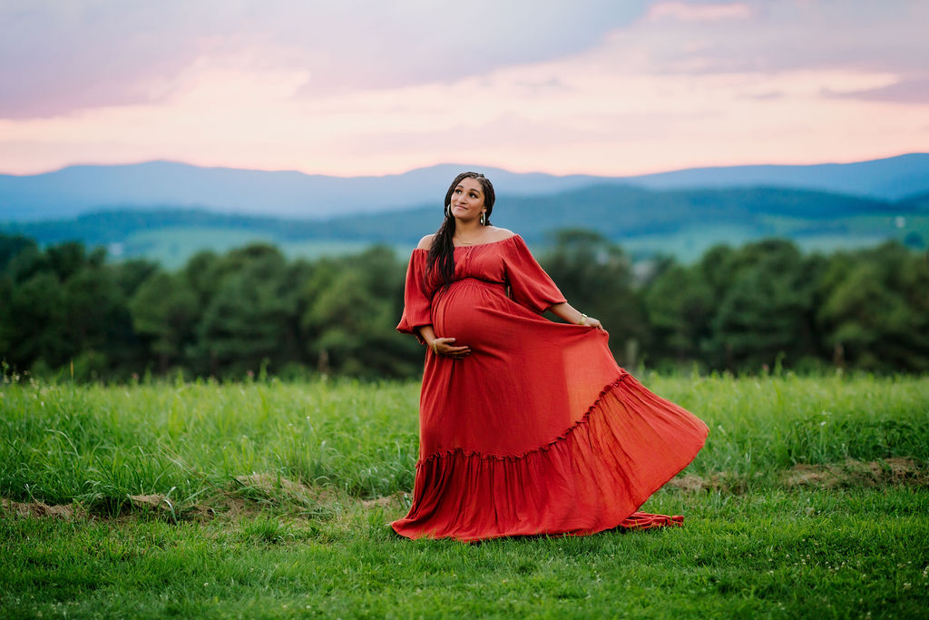 A mother to be dances in a grassy field at sunset while twirling her red maternity gown queen city birth services