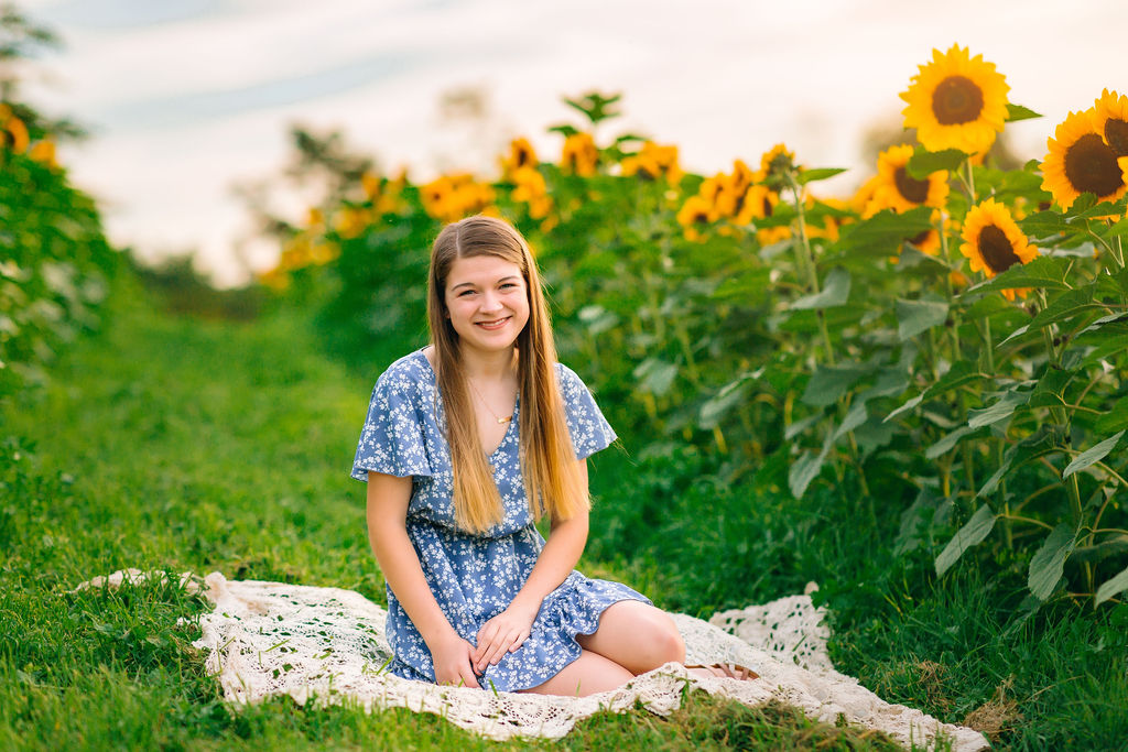 A young girl sits on a lace blanket in a field of sunflowers wearing a blue dress rocktown dental