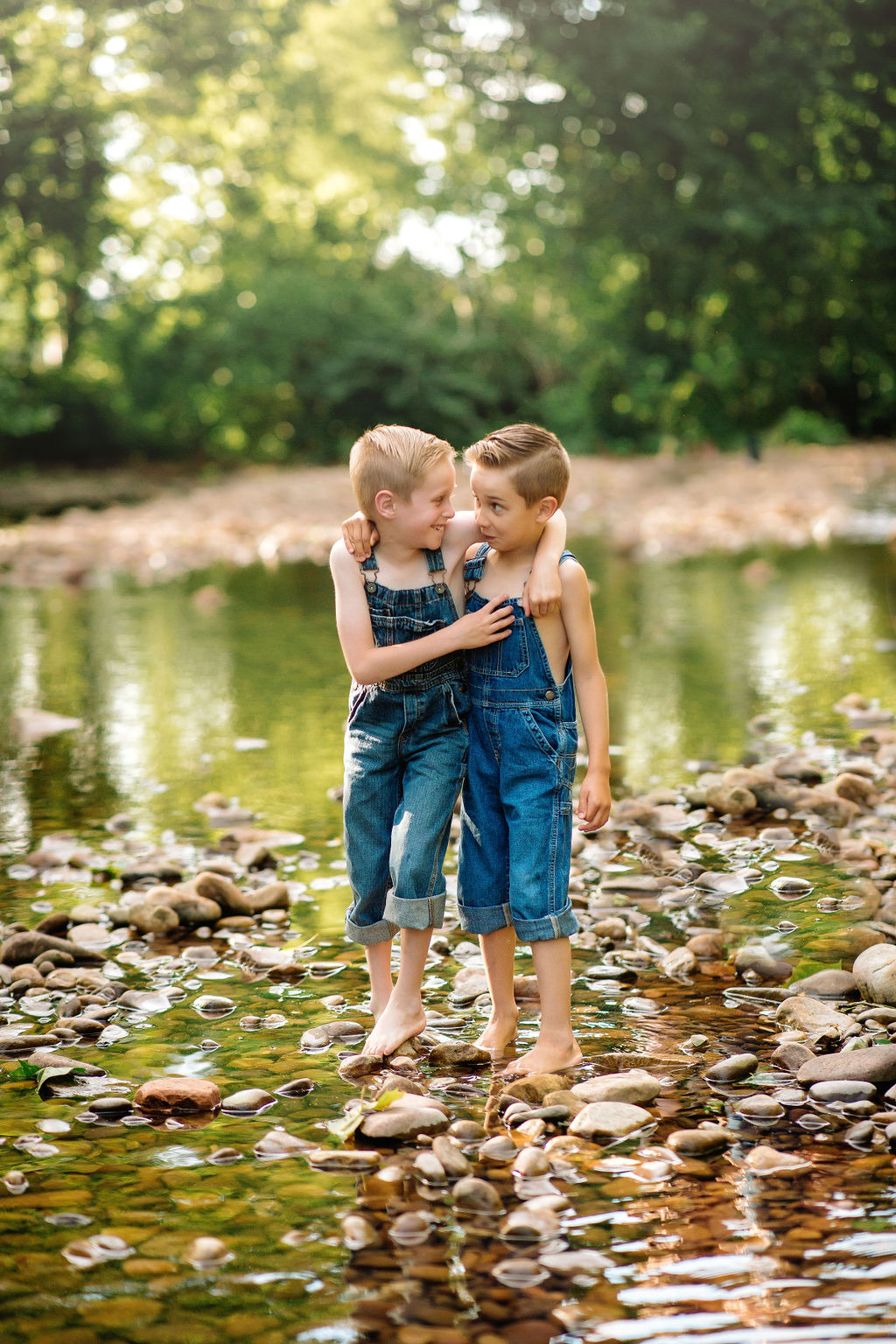 Two brothers wearing blue overalls hang onto each other while walking through a rocky stream