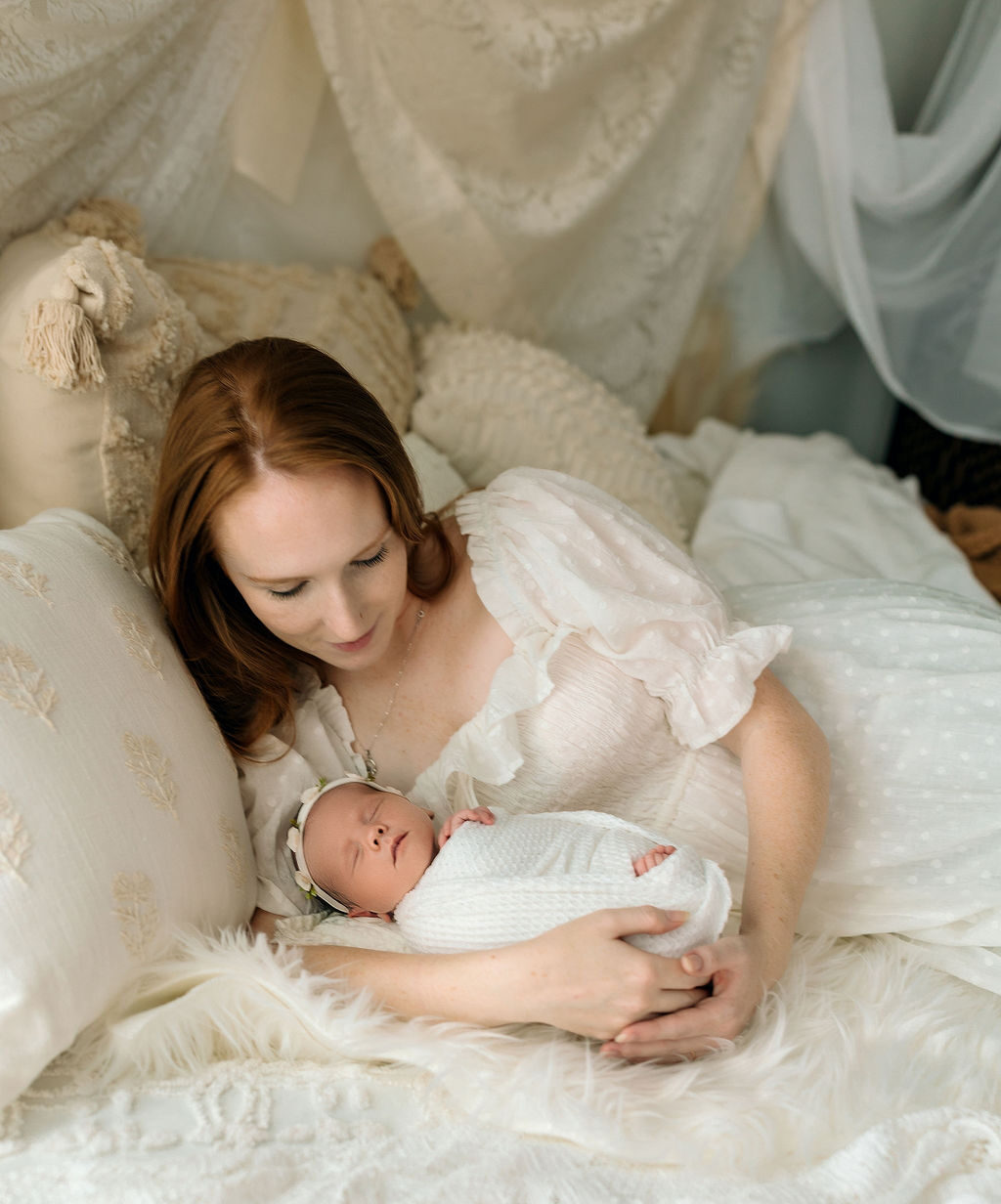 A mother in a white dress cuddles with her sleeping newborn baby on a bed virginia fertility and ivf