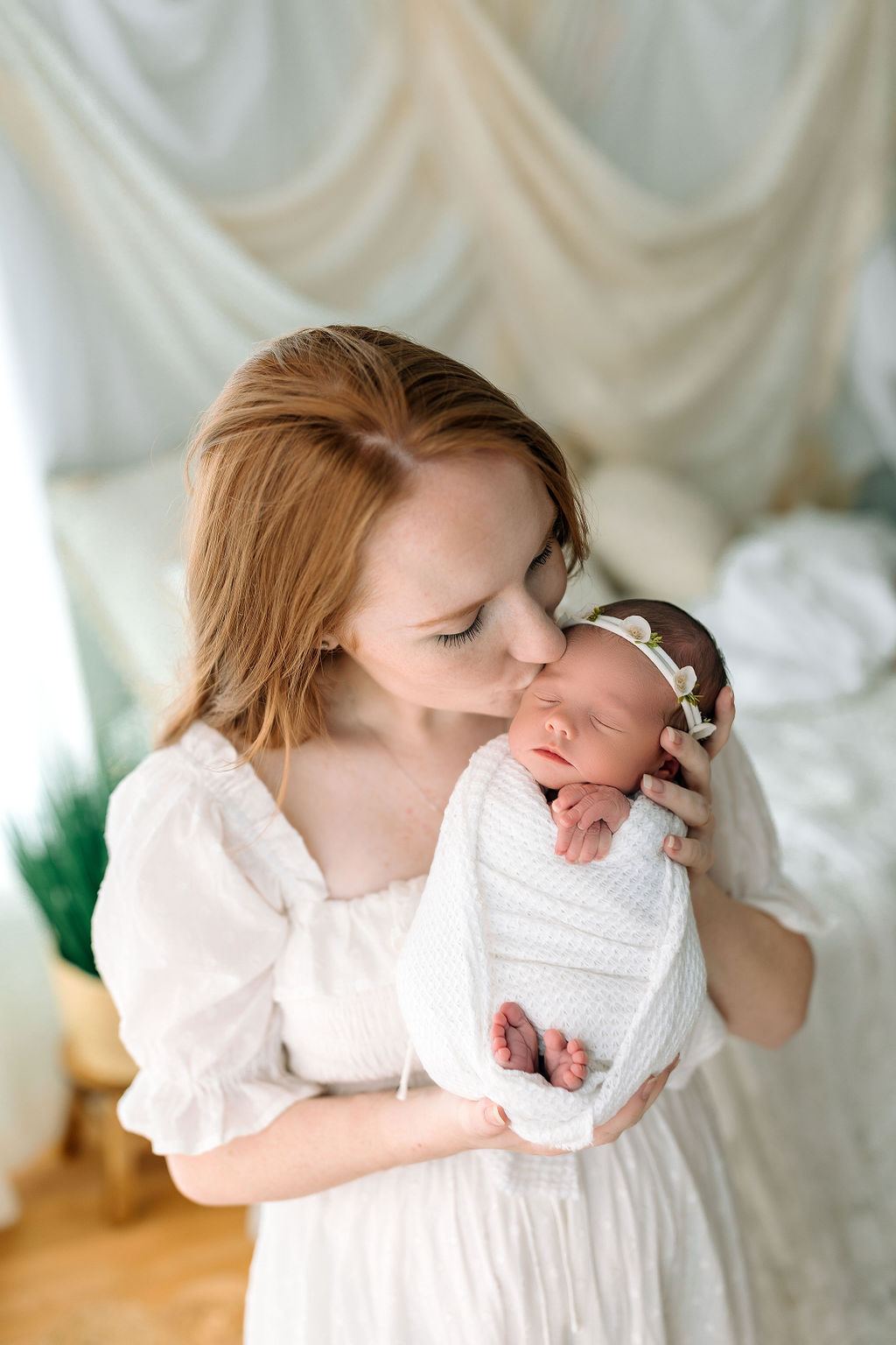 A new mother in a white dress kisses the cheek of her sleeping newborn baby in a white swaddle virginia fertility and ivf