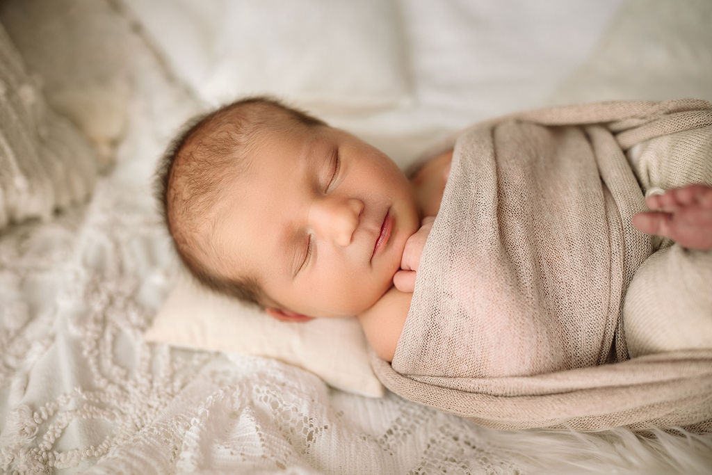A newborn baby sleeps in a brown cheesecloth swaddle on a lace blanket