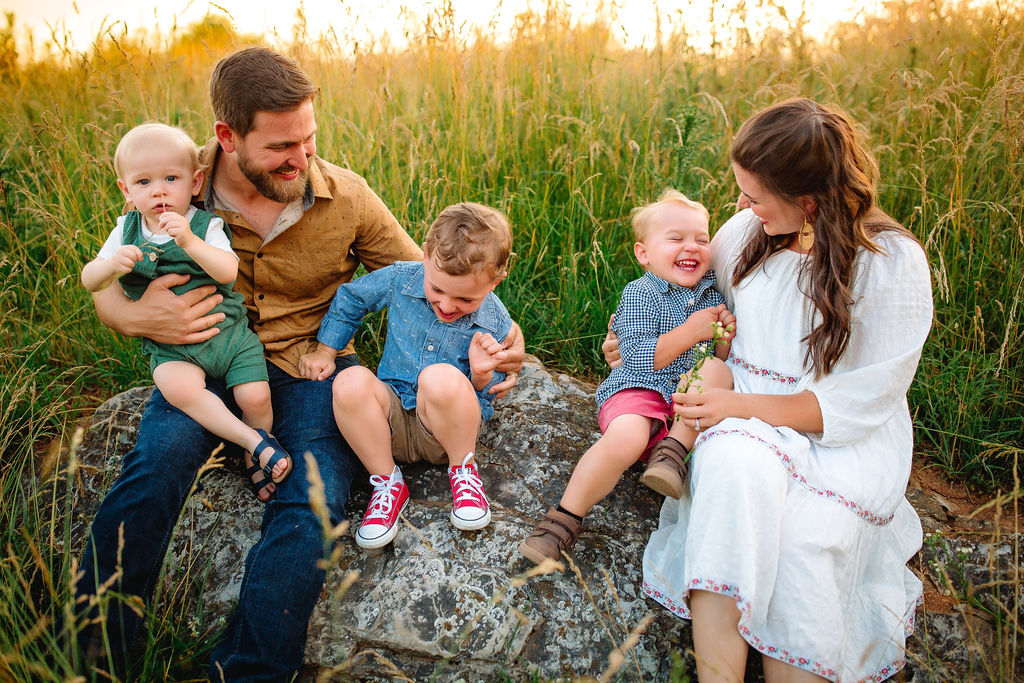 A mom and dad tickle their three children while sitting on a large rock in a field of tall grass