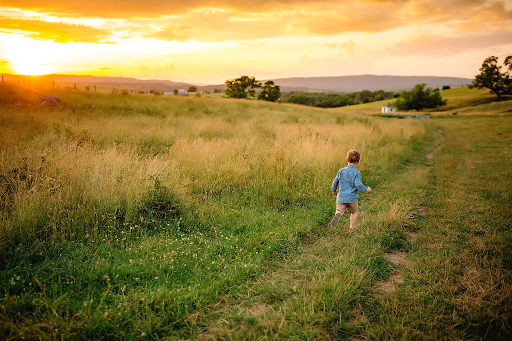 A young boy in a blue long sleeve shirt runs down a grassy path in a field at sunset back home on the farm