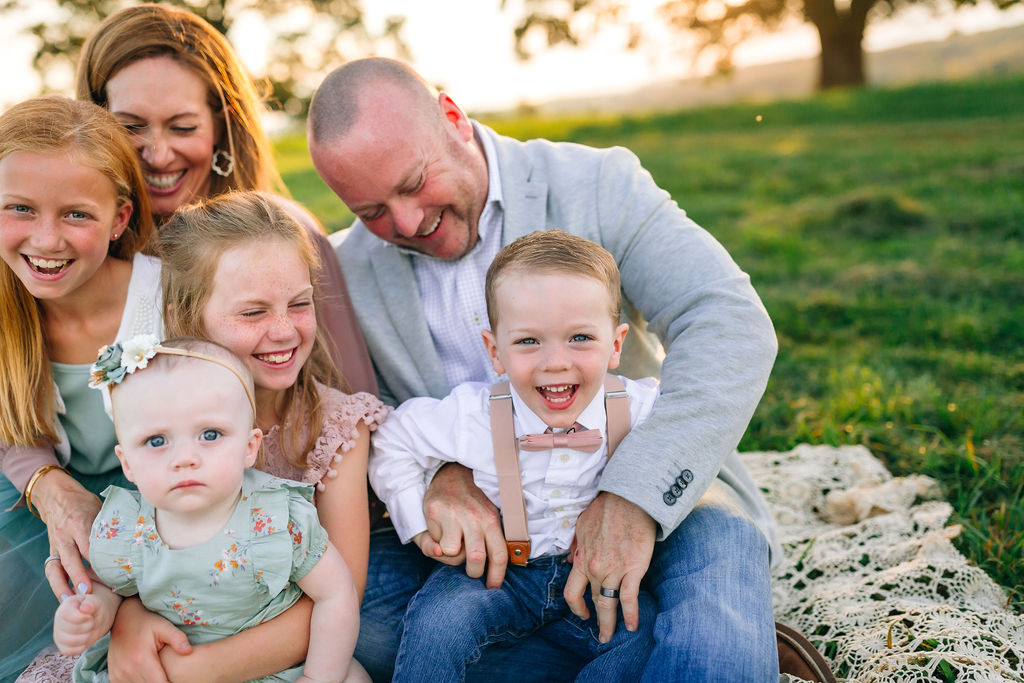 A family of six sit together on a lace blanket in a field tickling and playing with each other blue ridge music together