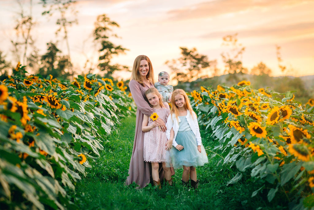 A mother stands in a field of sunflowers with an infant on her hip and two daughters leaning into her blue ridge music together