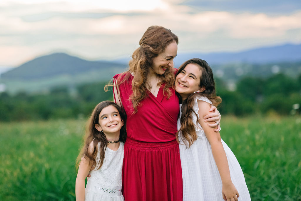 A mother in a red dress hugs her two daughters in a field wearing white dresses