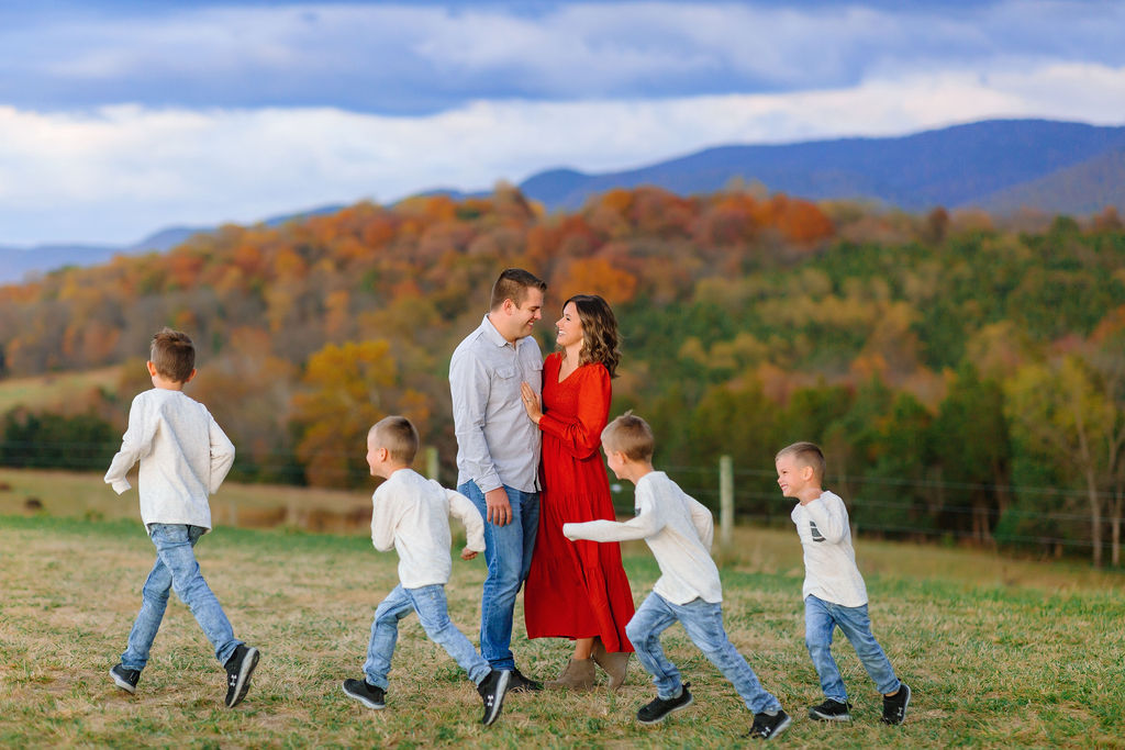 A mom and dad hug while their four sons runs circles around them in a grassy field