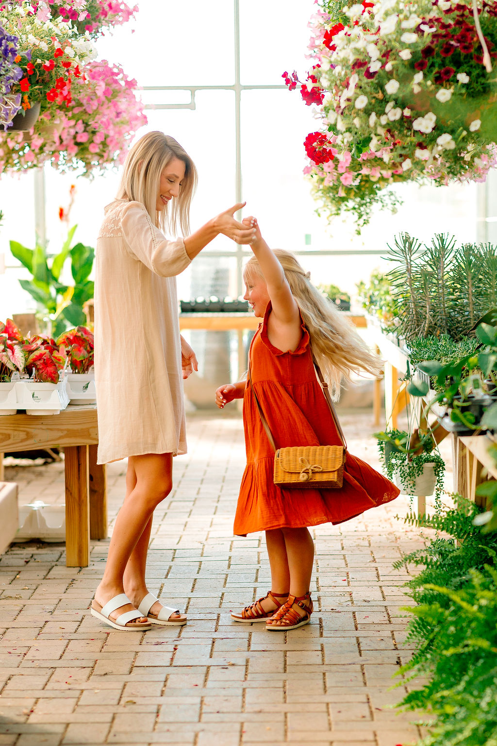 A mom dances with her young daughter in a greenhouse full of hanging flowers