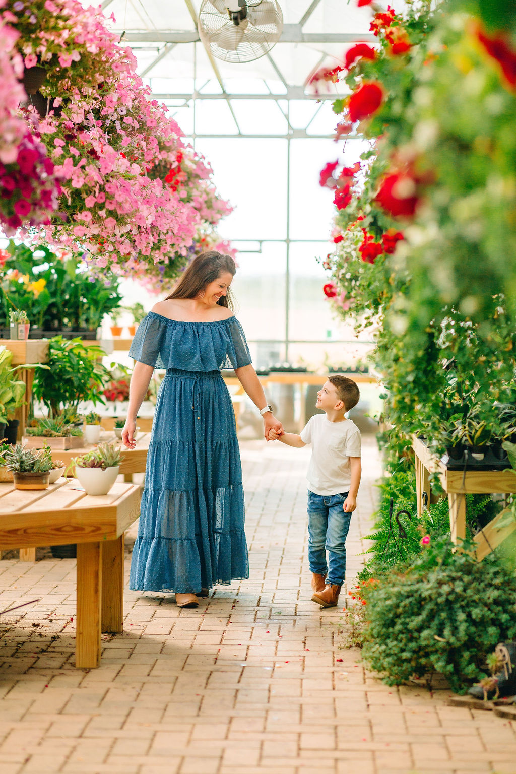 A mother and her young son walk through a greenhouse full of flowers holding hands