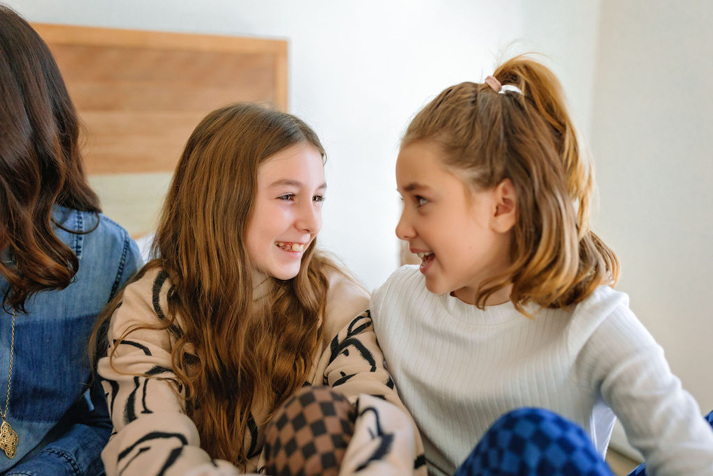 Two young sisters in sweaters sit on a bed looking and smiling at each other indoor things to do in winchester va